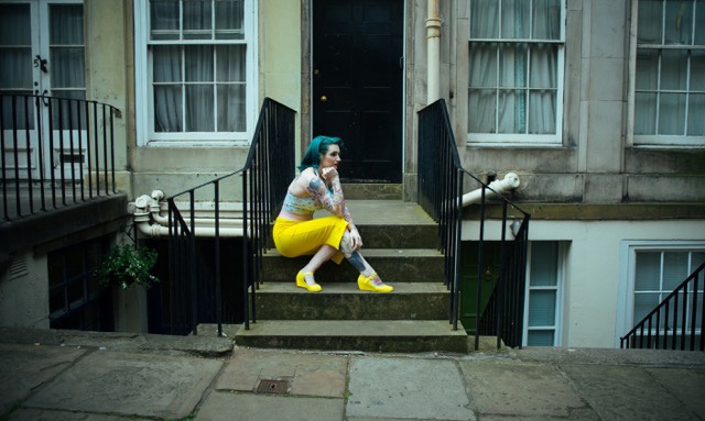 Miss Kelly Bakewell with green hair wearing a yellow pencil skirt sits on a stone stair
