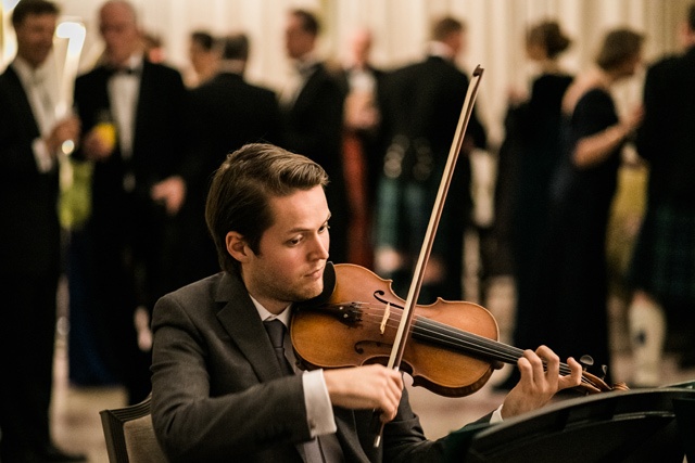 a young man sits playing the violin in a suit at a violin and piano recital