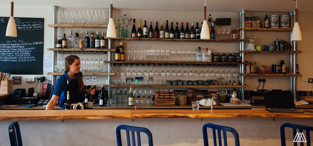 a young woman stands behind a bar with shelfs lined with wine bottles