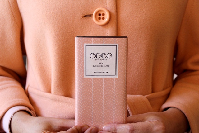 a woman in a peach coloured coat stands holding a peach coloured bar of chocolate