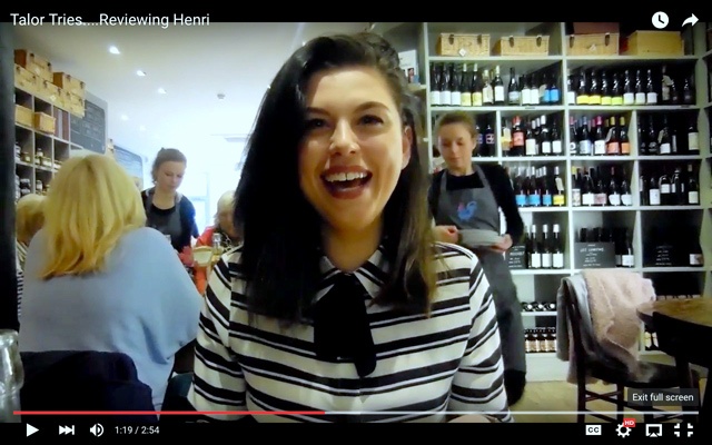 a young woman with shoulder length brunette hair wearing a black and white striped top sits in a busy cafe called Henris in Stockbridge