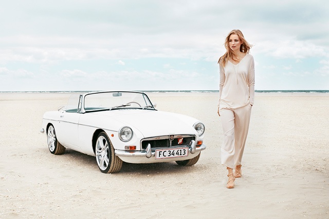 a slim female model with long blonde hair walks on a beach next to a white vintage open top sports car