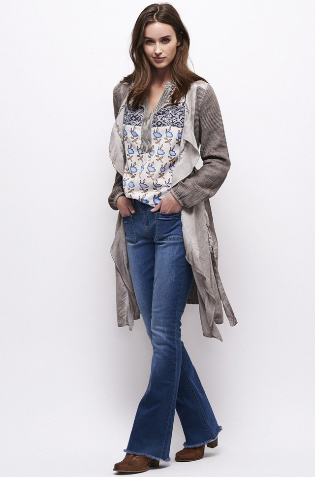 a slim female model with long brunette hair stands in a long cardigan and blue jeans