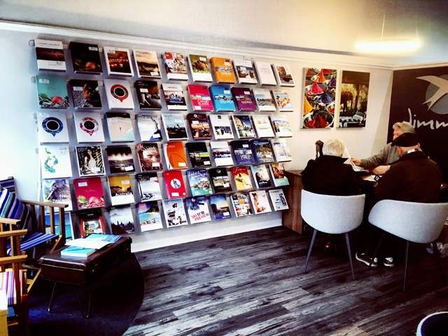 an elderly man and woman sit at a desk talking to a travel agent with rows of travel brochures on the walls
