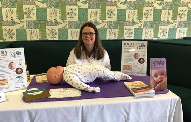 a woman in glasses sits behind a table with a toy baby on it at an Infant Massage Class