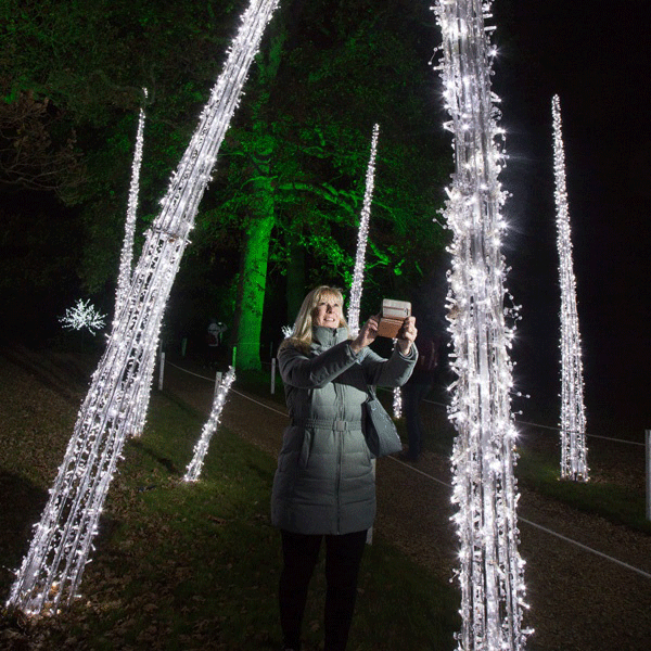 a woman in a large winter coat takes photographs at night with her phone of large oversized illuminated Christmas icicles
