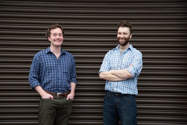 two young men wearing checked shirts stand in front of a brown corrugated garage door