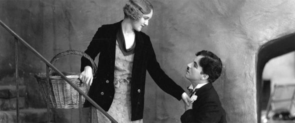 a woman carrying a wicker basket stands on a stair above a man while he holds her hand to his chest