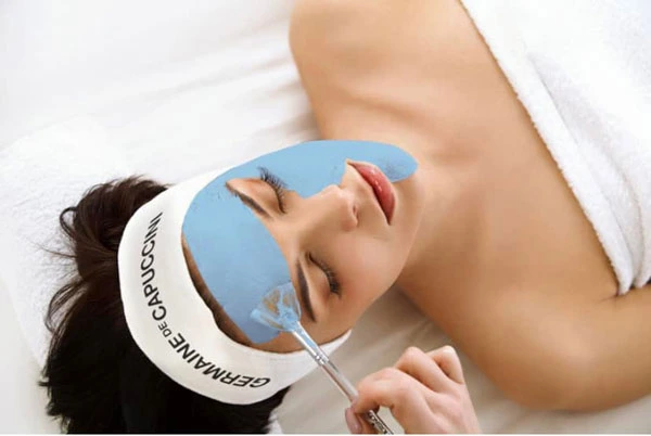 A brunette woman lying on a table wrapped in a white towel receiving a facial treatment