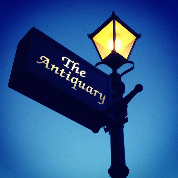 An illuminated old fashioned lamp post with a sign attached for The Antiquary Bar in Stockbridge, Edinburgh